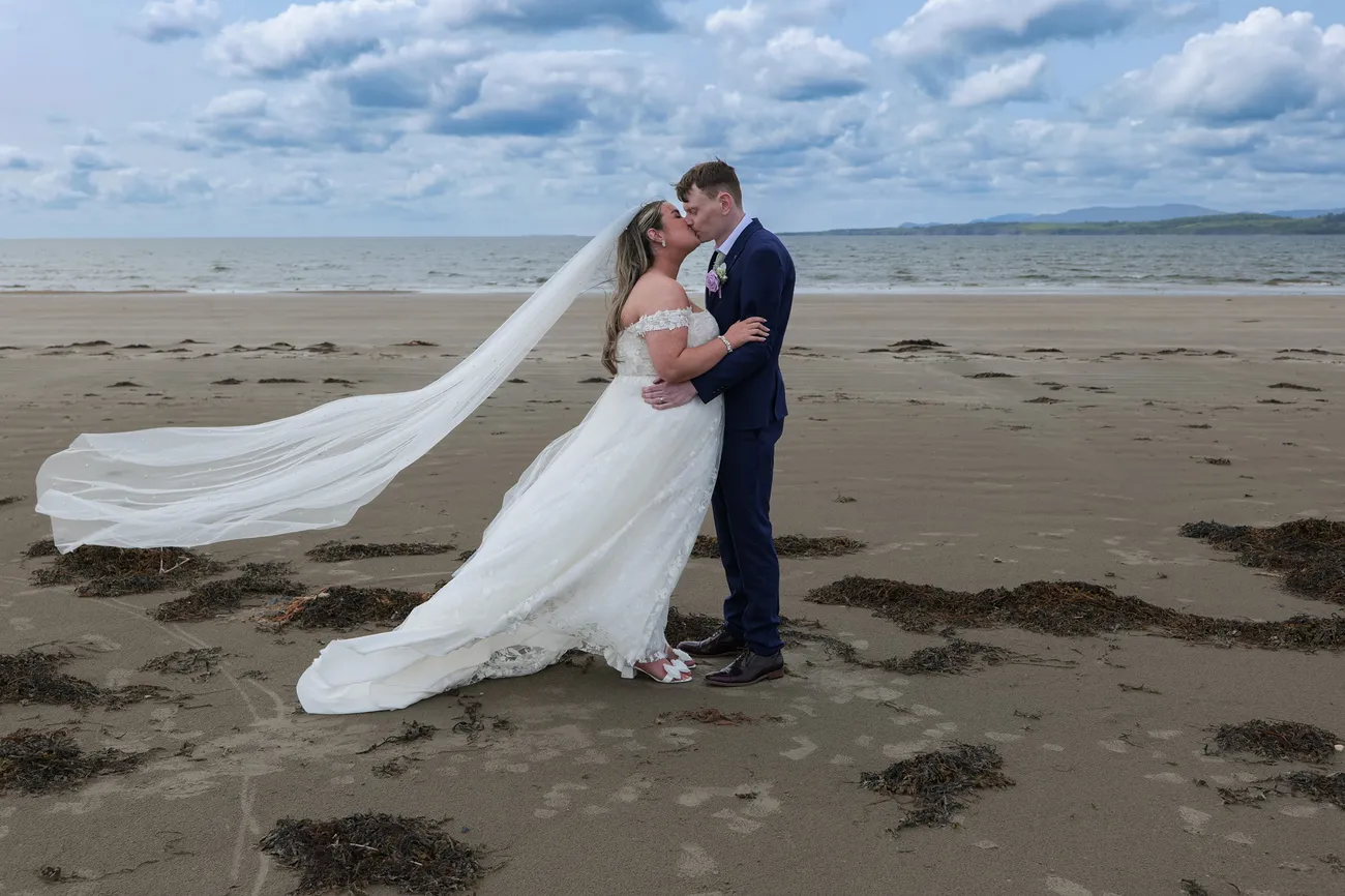Bride and groom kissing on a beach with a windswept veil