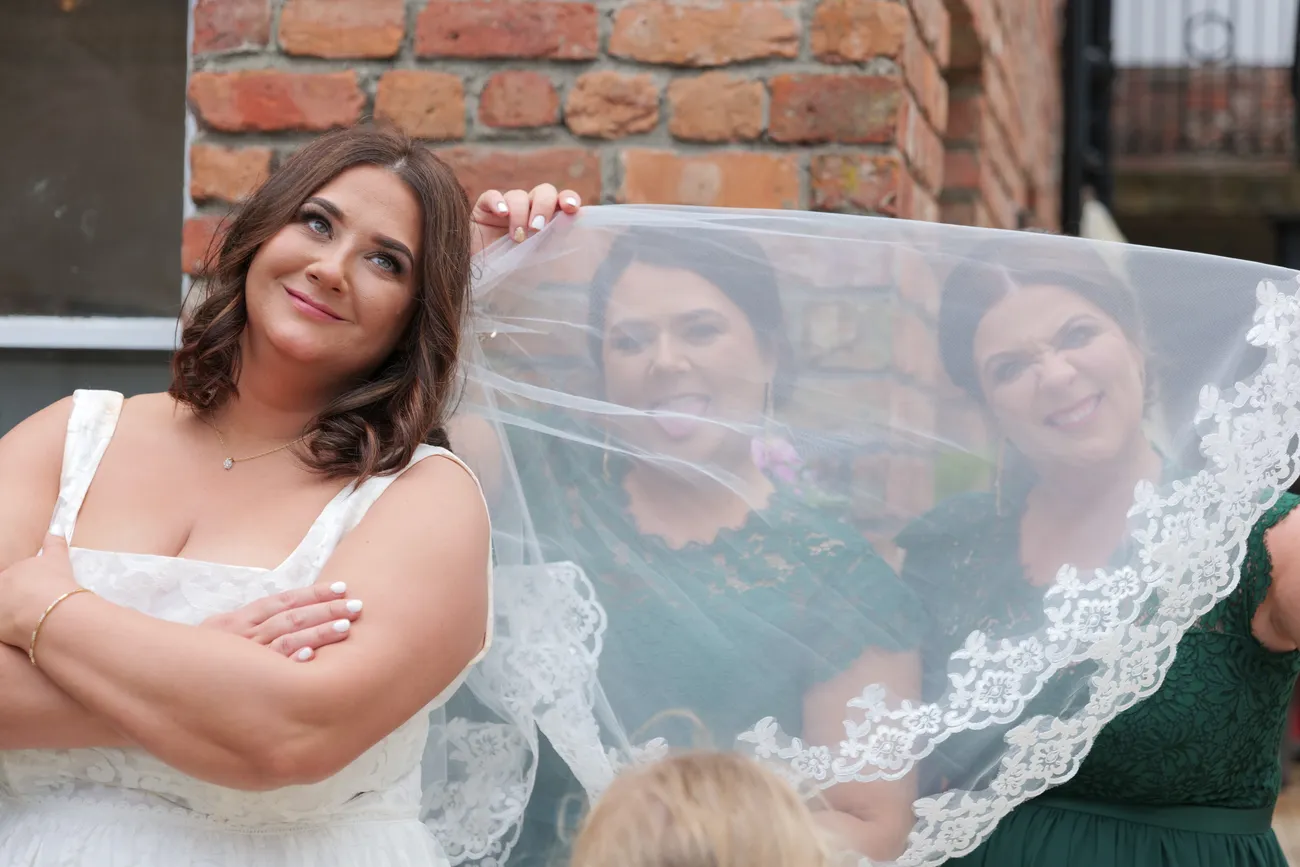 Bride under a veil beside a brick wall with bridesmaids behind