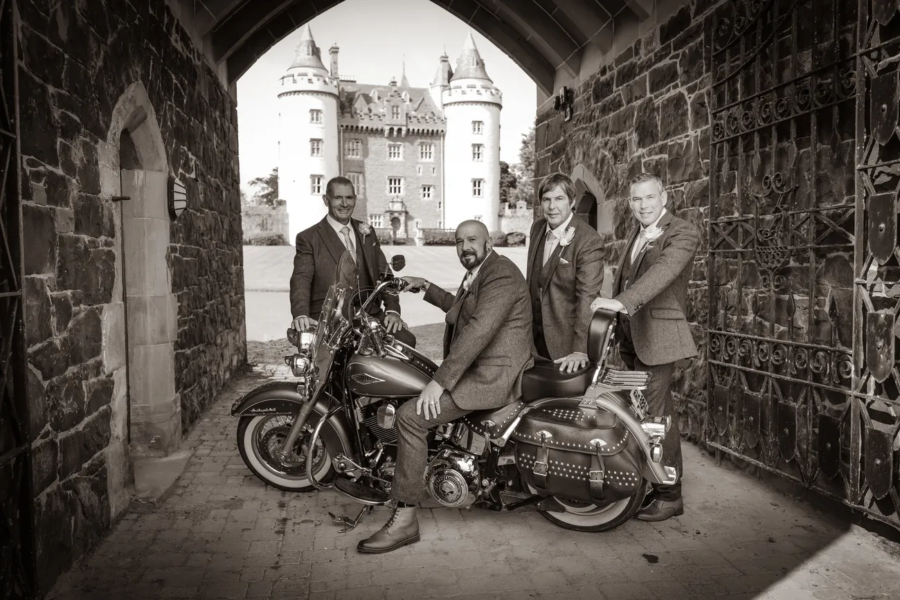 Groomsmen posed with a motorbike under a castle arch