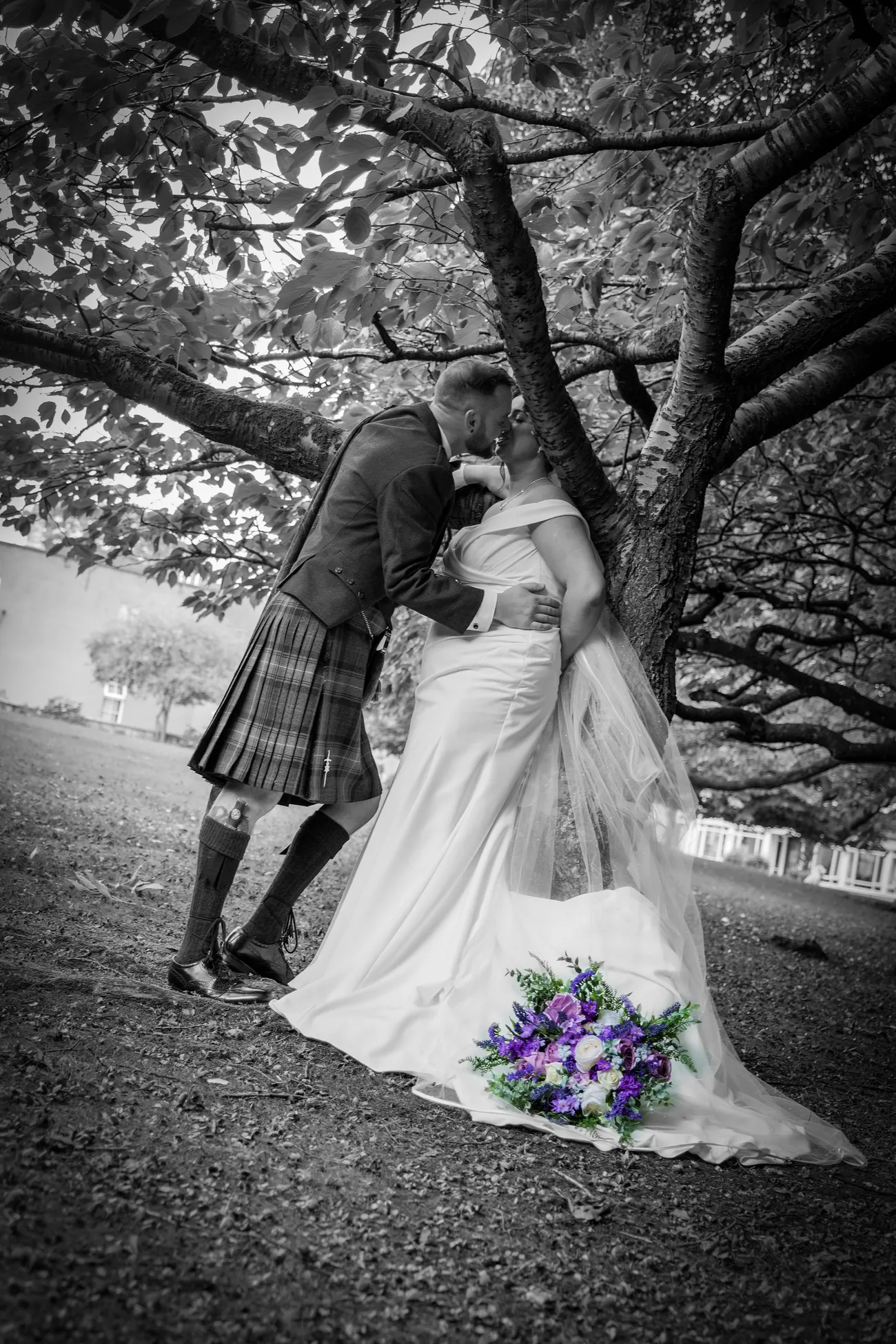 Kilted groom kissing the bride beneath a tree in black and white